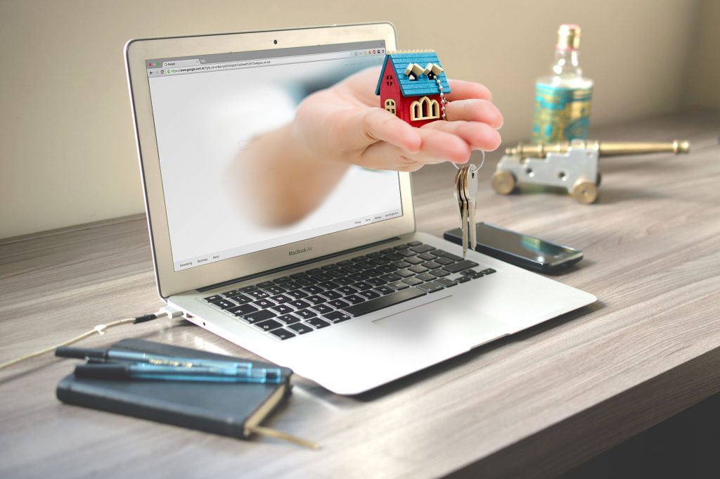 um notebook com uma mão saindo da tela segurando uma casa em miniatura